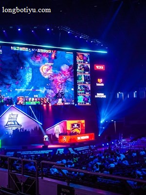 A large esports arena featuring a massive screen displaying a video game, surrounded by sponsor logos and team names. The auditorium is filled with spectators watching the gaming event. Neon blue and purple lights illuminate the stage, creating a vibrant atmosphere.