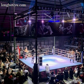 A crowded indoor arena with a boxing ring at the center, surrounded by an audience seated at tables. The scene is illuminated by overhead lights, casting a vibrant glow over the ring where two fighters are preparing for a match. Spectators are engaged, capturing the event with cameras and enjoying the lively atmosphere.